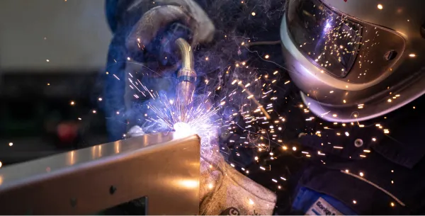 Ein Schweißer mit Schutzhelm und Handschuhen bei der Arbeit, während helle Funken und Rauch beim Schweißen eines Metallrahmens entstehen.