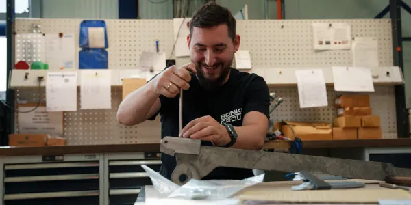 Ein lächelnder Mitarbeiter montiert ein Metallbauteil mit einem Inbusschlüssel an einer Werkbank in einer Werkstatt.